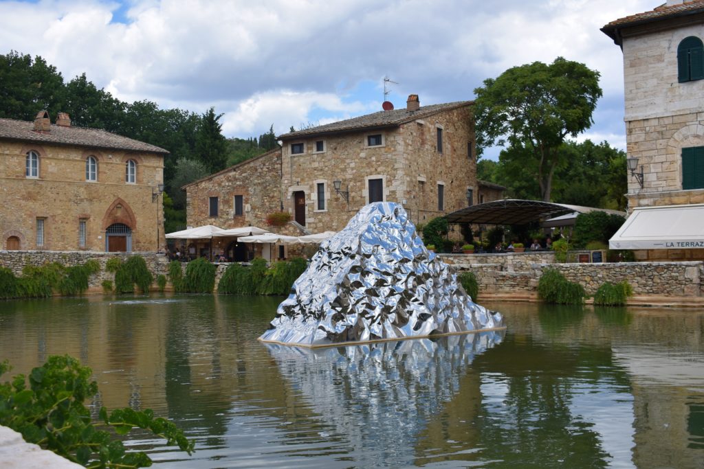 Surrounded by small medieval houses A pool filled with water with the silver sculpture standing in the middle of it 