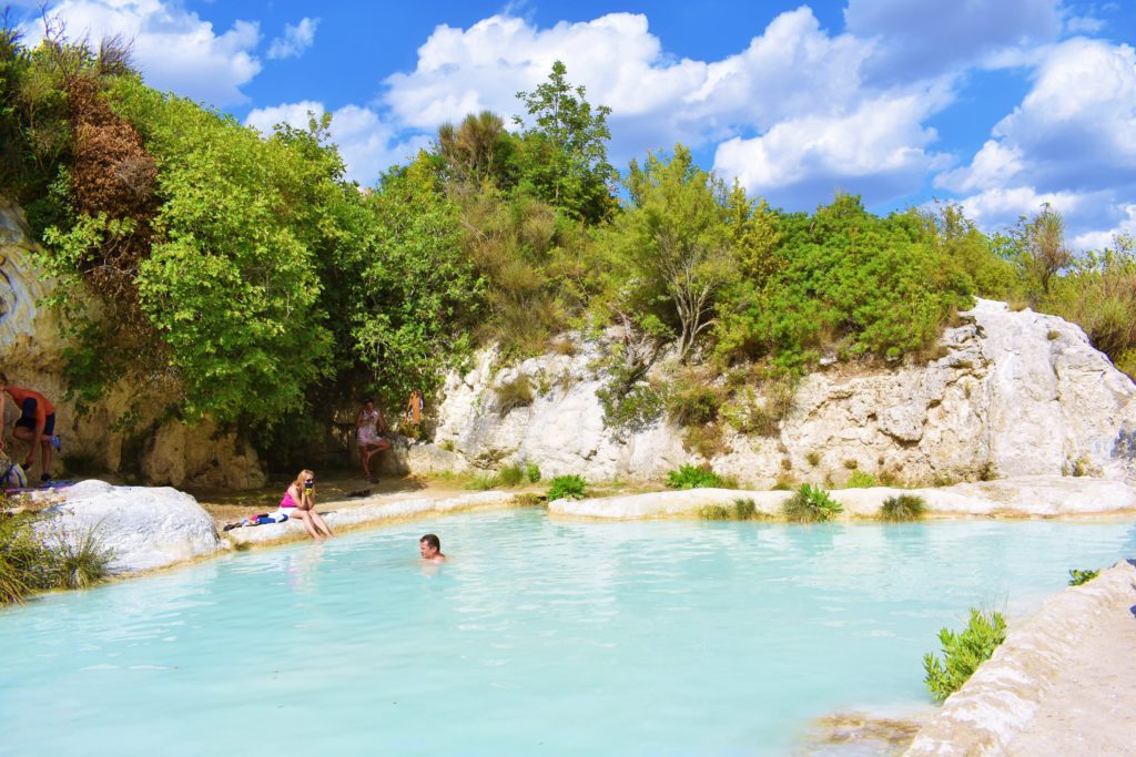 People having bath in an natural outdoor pool filled with blue thermal water among white stones and trees. 